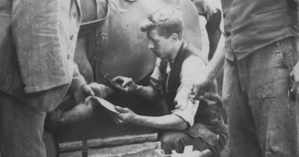 Josef Rauch kneels and works on one of the horse's hooves while another person holds the horse's hoof. A third person watches Josef at work.