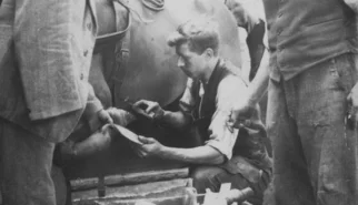 Josef Rauch arbeitet kniend an einem der Hufe eines Pferdes, während eine andere Person den Huf des Pferdes hält. Eine Dritte Person beobachtet Josef bei der Arbeit.