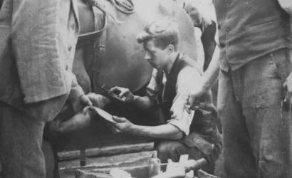 Josef Rauch kneels and works on one of the horse's hooves while another person holds the horse's hoof. A third person watches Josef at work.